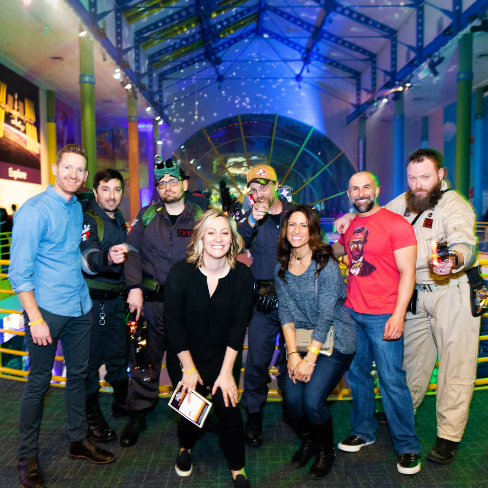 A group of adults posing with Ghostbusters in the Sunburst Atrium during Museum by Moonlight
